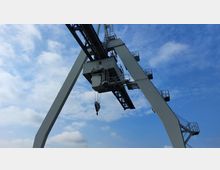 Ein Industriekran mit großen, weißen Metallstreben und einer Kabine in der Mitte, umgeben von Gerüsten und Treppen, vor einem blauen Himmel mit wenigen Wolken.