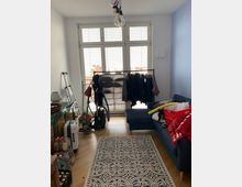 Ein Wohnzimmer mit hellem Holzboden, großen Fenstern mit weißen Fensterläden und einer blauen Wand. Im Raum befinden sich ein Sofa, ein Teppich mit geometrischem Muster, ein Wäscheständer mit Kleidungsstücken, Haushaltsgeräte wie ein Staubsauger, sowie diverse Gegenstände, darunter Kartons und Getränkeflaschen.