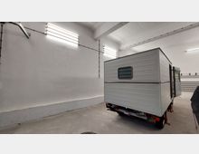 Innenraum einer großen, weißen Halle mit hohen Decken und minimaler Einrichtung. Im Vordergrund ein kleiner, grauer Anhänger mit Fenster, stehend auf Stützen; an den Wänden sind neonbeleuchtete Lampen und Kabel zu sehen.