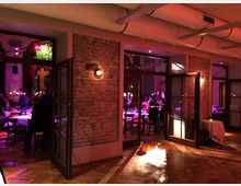Innenraum eines stilvoll beleuchteten Restaurants oder Veranstaltungsraums mit freiliegenden Backsteinwänden, Glastüren und Parkettboden. Im Hintergrund befinden sich gedeckte Tische und Gäste in einem gemütlichen, von warmem Licht erfüllten Ambiente.