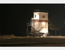 Ein einfacher Kontrollturm auf einem Flugplatz bei Nacht, beleuchtet durch künstliches Licht, mit einer kleinen Propellermaschine im Vordergrund und einem Baum am rechten Bildrand.