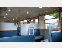 Innenraum eines Zugwagons mit blauen Sitzen und weiß-grauen Trennwänden. Große Fenster mit Holzumrahmung bieten Blick nach draußen, wo ein vorbeifahrender Zug und grüne Vegetation zu sehen sind.