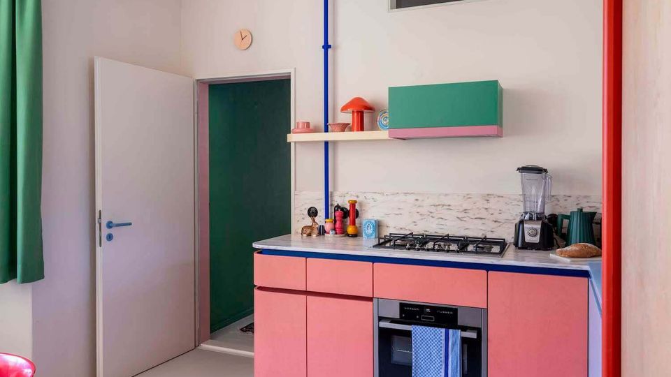 Colorful modern kitchen with pink lower cabinets, a marble-look countertop, and a built-in oven beneath a gas cooktop. White walls feature open shelves and a green wall cabinet, with exposed red and blue pipes, a small high window, and an open door leading to another room.