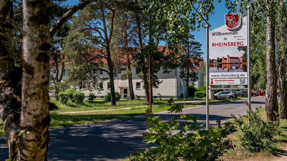Straßenszene am Ortseingang mit einem großen Willkommensschild „Rheinsberg“ neben einer zweispurigen Straße und Gehweg. Im Hintergrund stehen mehrstöckige Wohngebäude mit roten Ziegeldächern, umgeben von Bäumen und Grünflächen.