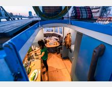 Innenraum eines Hausbootes mit Holzboden und blauen Wänden. Im Vordergrund eine kleine Küche mit Spüle und Gemüse auf der Arbeitsfläche; im Hintergrund eine Sitzecke mit Personen an einem Tisch und Blick auf das Wasser durch ein halbrundes Panoramafenster.