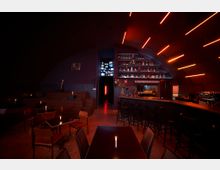 Elegante Bar im Innenbereich mit dunkler Wandgestaltung, moderner Beleuchtung durch rote Neonröhren und einer Theke mit Regalwänden voller Flaschen im Hintergrund. Der Raum bietet Sitzgelegenheiten mit einfachen Tischen und Stühlen sowie eine Bühne mit Stufen und Kerzenlicht.