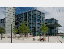 Moderne Glasfassade eines Bahnhofsgebäudes mit mehreren rechteckigen Flügeln und einem geschwungenen Glasdach über dem Eingangsbereich. Vor dem Gebäude befinden sich ein Platz mit Bänken, Bäumen, Laternen und geparkten Taxis.