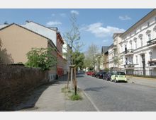 Straßenszene in einem Wohngebiet mit gepflastertem Gehweg und Straße, gesäumt von mehrstöckigen Altbauten mit Balkonen auf der rechten Seite. Links ist eine Ziegelmauer, daneben moderne Häuser in Pastellfarben sowie junge Bäume entlang des Bürgersteigs.