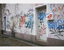 Außenwand eines Gebäudes mit weißem Putz, vollständig mit buntem Graffiti und Tags bedeckt; im Vordergrund befinden sich eine Tür und ein Briefkasten, daneben ein gepflasterter Gehweg.