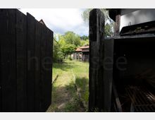 Blick durch ein geöffnetes, dunkel gestrichenes Holztor auf einen schmalen, grasbewachsenen Gartenweg mit niedrigen Schienen im Boden. Im Hintergrund stehen Bäume, Zäune und ein kleines Gebäude mit rotem Dach.