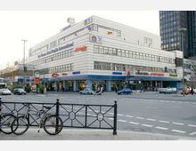 Eckgebäude mit weißer Fassade in einer städtischen Umgebung, das ein Hotel ("Best Western Hotel Steglitz International") und mehrere Geschäfte im Erdgeschoss beherbergt, darunter eine Apotheke und ein Schuhladen. Im Vordergrund eine Straßenkreuzung mit Autos, Fußgängern und geparkten Fahrrädern.