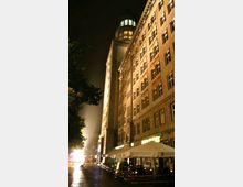 Nachtszene einer Stadtstraße mit einem beleuchteten historischen Gebäude im Zentrum, das sich durch hohe Fenster und eine auffällige Turmspitze mit grünlicher Beleuchtung auszeichnet. Im Vordergrund befinden sich Außensitzplätze eines Restaurants mit weißen Sonnenschirmen.