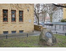 Eine Außenansicht einer gelben Backsteinfassade mit mehreren Fenstern, daneben ein gepflasterter Gehweg und eine Straße im Hintergrund. Im Vordergrund steht ein großer Findling mit einer runden Gedenkplakette, umgeben von einer Grünfläche und niedrigen Metallabsperrungen.