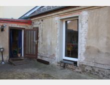 Außenansicht eines renovierungsbedürftigen Gebäudes mit verputzter und teilweise freiliegender Ziegelwand. Zu sehen sind eine braune Tür mit Glaseinsatz, ein Fenster mit weißem Rahmen und ein kleiner Außenbereich mit Betonboden und bräunlichen Flecken.