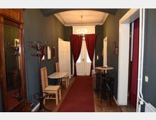 Ein langer, schmaler Flur mit dunkelgrauen Wänden, rotem Teppich und hölzernem Boden. Auf der linken Seite stehen ein antiker Spiegel, eine Garderobe und Holzmöbel, auf der rechten Seite befinden sich ein kleiner Tisch mit Barhockern und gerahmte Bilder; im Hintergrund weiße Doppeltüren mit roten Vorhängen und ein Kronleuchter an der Decke.