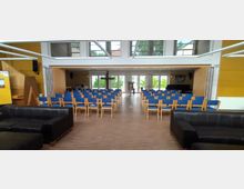 Ein moderner Gemeindesaal mit blauen Stühlen in Reihen angeordnet, einem Holzkreuz und einem Pult im Vordergrund der Bühne. Große Fenster im Hintergrund lassen Tageslicht herein, und schwarze Sofas stehen im Vordergrund des Raumes.