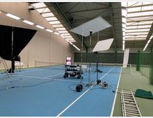 Ein Innenraum einer Tennishalle mit blauen Spielfeldern und grünen Begrenzungen. Im Vordergrund sind Kamera- und Beleuchtungsequipment sowie Kabel und Leitern für eine Film- oder Fotosession aufgebaut.