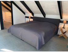 Schlafzimmer unter einer Dachschräge mit sichtbaren schwarzen Holzbalken. Im Raum steht ein großes Doppelbett mit dunkler Tagesdecke, daneben befinden sich ein Nachttisch mit Lampe sowie eine offene Tür, die zu einem angrenzenden Raum mit Regalen führt.