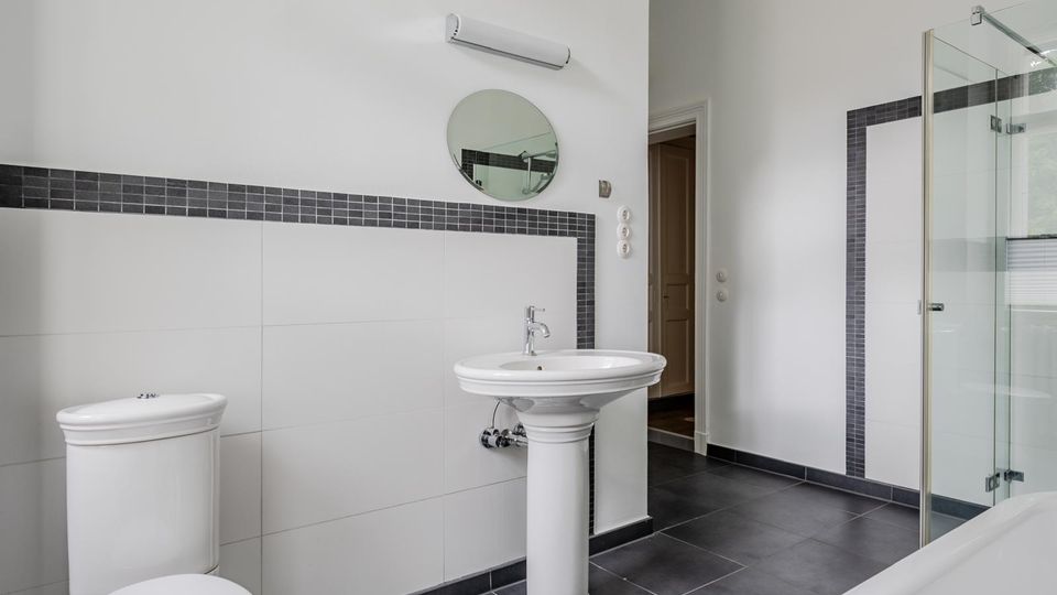 Modernes Badezimmer mit weißen Wandfliesen und dunklem Fliesenboden, ausgestattet mit Standwaschbecken, Toilette und rundem Spiegel unter einer Wandleuchte. Rechts ist eine Badewanne mit Glasduschwand sowie ein Fenster mit Sicht ins Grüne zu sehen.