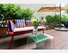 Moderne Terrasse mit Holzboden, auf der eine rote Gartenbank mit gestreiften Kissen und ein kleiner Tisch mit einer Vase voller Blumen stehen. Im Hintergrund befinden sich grüne Pflanzen, Hochbeete und ein großer Sonnenschirm.