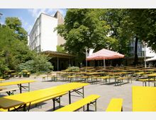 Ein Biergarten mit gelben Holzbänken und Tischen auf einem gepflasterten Platz, umgeben von Bäumen und Sträuchern. Im Hintergrund befinden sich ein weißes Gebäude, eine überdachte Terrasse und ein rot-weiß gestreifter Sonnenschirm.