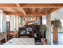 Ein rustikaler Innenraum mit Holzbalkendecke, einer Holztreppe im Hintergrund und einem Bücherregal, das mit Büchern und Dekorationen gefüllt ist. Im Vordergrund steht ein Tisch mit Stühlen, und Pflanzen schmücken den Raum, während Fenster natürliches Licht hereinlassen.