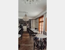 Eleganter Café- oder Restaurantinnenraum mit Kronleuchtern, dunklen Holzstühlen und weiß gedeckten Tischen auf Fischgrätparkett. Rechts sorgen hohe Holzfenster und klassisch dekorierte Wandpaneele in Grautönen für Tageslicht und einen historischen Ambientecharakter.