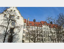 Großes historisches Gebäude mit heller Putzfassade und rotem Ziegeldach, geprägt von geschwungenen Giebeln, vielen Fenstern und kleinen Dachaufbauten mit Türmchen. Im Vordergrund stehen kahle Bäume vor der Fassade unter blauem Himmel.
