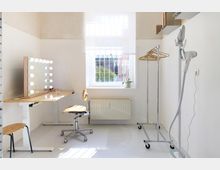 Heller Ankleideraum mit einem Schminktisch mit beleuchtetem Spiegel, einem Stuhl auf Rollen, einem Kleiderständer und einem Dampfbügler. Ein Fenster mit Fenstergitter lässt Tageslicht herein, und eine kleine Pflanze steht auf dem Fensterbrett.