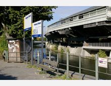 Uferbereich eines Kanals mit einer erhöhten Stahlkonstruktion einer Bahnstation im Hintergrund. Im Vordergrund befinden sich Schilder mit der Aufschrift „Reederei Riedel“, eine Sitzbank, sowie ein kleiner beleuchteter Bereich mit Graffiti.
