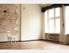 Ein leerer Raum mit einem hellen, hölzernen Fischgrätenboden. Eine Wand zeigt freiliegendes Mauerwerk mit einer teilweisen Putzschicht, daneben ein großes Fenster mit schwarzem Vorhang und darunter freiliegende Heizungsrohre. Ein einzelner weißer Stuhl steht an der Wand.