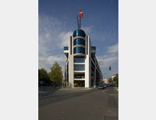 Eckgebäude mit moderner Architektur und weißer Fassade, Akzente aus dunkelblauen Glasflächen und einem spitzen Aufsatz mit roter SPD-Flagge; vor dem Gebäude verläuft eine Straße mit Bäumen und parkenden Fahrzeugen im Umfeld.