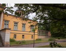 Gelbes, klassizistisches Gebäude mit großen Fenstern und umlaufenden Zierelementen, flankiert von einer offenen Säulengalerie auf der rechten Seite. Im Vordergrund befinden sich ein Weg, Rasenflächen und Bäume.