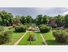 Parkanlage mit symmetrischen Blumenbeeten, Rasenflächen und Gehwegen, die zu einem zentralen, runden Springbrunnen führen. Im Hintergrund sind Bäume und rote Backsteingebäude zu sehen.