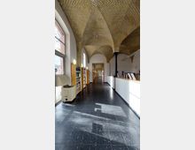 Langer Bibliotheksgang mit gewölbter Deckenkonstruktion aus gelbem Ziegelstein, schwarzen Bodenfliesen und hohen Fenstern auf der linken Seite. Rechts befinden sich Lesetische, und auf der linken Seite stehen Bücherregale.