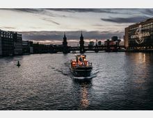Ein Boot fährt auf einem Fluss in Richtung Kamera, flankiert von modernen Gebäuden und der beleuchteten Oberbaumbrücke im Hintergrund. Rechts im Bild ist ein Gebäude mit dem Schriftzug „Universal“ zu sehen, der Himmel ist wolkenbedeckt und zeigt eine Dämmerungsstimmung.