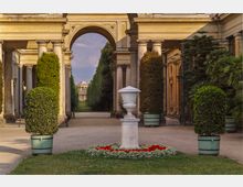 Eine gepflegte Gartenanlage mit symmetrischen Hecken und Buchsbaumkübeln in Töpfen. Im Vordergrund eine weiße Steinvase auf einem Sockel, umgeben von roten Blumen, und im Hintergrund ein monumentaler Torbogen, der den Blick auf ein prunkvolles Gebäude mit Kuppel lenkt.