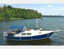 Ein kleines Motorboot mit blau-weißer Lackierung und einem blauen Sonnendach liegt vor Anker auf einem See, umgeben von dichten grünen Bäumen am Ufer. Im Hintergrund sind weitere Boote und der bewaldete Horizont zu sehen.