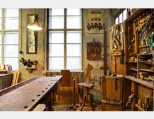 Werkstatt mit rustikalem Holztisch, Werkzeugwand und Regalen; an den teils verputzten Wänden hängen Werkzeuge und Zeichnungen, während Licht durch große Sprossenfenster einfällt. Im Raum stehen ein Holzstuhl sowie verschiedene Holzbretter und Möbelteile verteilt.