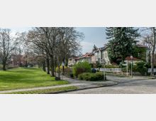 Parkähnliche Stadtlandschaft mit einer Grünfläche links, gesäumt von kahlen Bäumen, sowie einer gepflasterten Straße rechts, an der Reihenhäuser mit Vorgärten und Zäunen stehen. Im Hintergrund verläuft ein Fußweg entlang der Grünfläche.