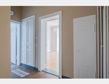 Heller Wohnungsflur mit beigefarbenen Wänden, weiß gerahmten Türen und Einbauschrank sowie sichtbaren Rohrleitungen an der rechten Wand. Durch eine offene Tür blickt man in einen weiteren Raum mit Parkettboden und Deckenleuchte.