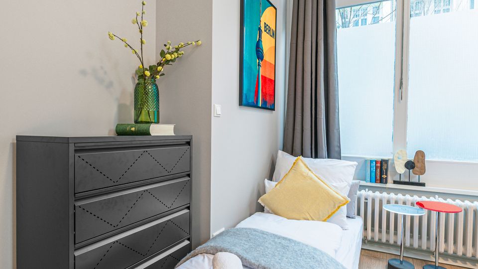 Schmales, modern eingerichtetes Schlafzimmer mit Einzelbett, schwarzer Kommode und hellem Holzboden. Große Fensterfront mit grauen Vorhängen und Heizkörper darunter, daneben ein kleines Beistelltisch-Set und ein farbiges Wandbild.