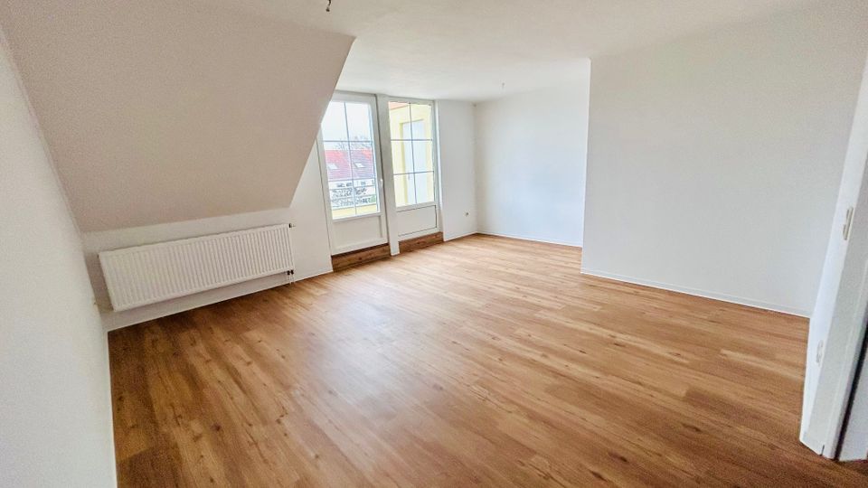 Leerer, heller Wohnraum mit Holzfußboden, weißen Wänden und einer Dachschräge. Große Balkontür-Fensterfront mit Geländerblick sowie ein Heizkörper an der linken Wand.