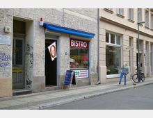 Straßenszene mit einem kleinen Bistro mit rotem Schild, einer beschrifteten Tafel und einer Hängemarke vor dem Eingang. Daneben befindet sich ein Schaufenster mit der Aufschrift „Schokogalerie“, ein Mann kehrt den Gehweg, und ein Fahrrad ist an einer Laterne angelehnt.