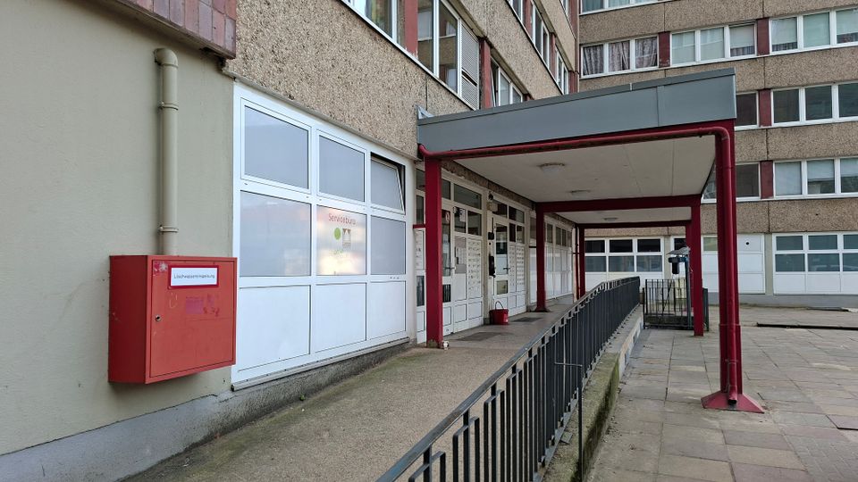 Eingangsbereich eines mehrstöckigen Wohn- oder Verwaltungsgebäudes mit überdachter Rampe und Metallgeländer, weißen Fenster- und Türfronten sowie einem roten Brandschutzkasten an der Außenwand. Der Zugang liegt in einem gepflasterten Innenhof mit sichtbaren Rohrleitungen und Stützen unter dem Vordach.