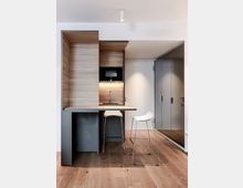 Moderne Kochnische mit Holzverkleidung und integriertem Waschbecken sowie Mikrowelle. Davor steht ein kleiner Tisch mit zwei weißen Barhockern, auf einem Holzboden; rechts ein Wandabschnitt mit Spiegeltüren.