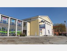 Gelbes Gebäude mit Säulenvorbau, das als "Friedrich-Wolf-Theater" beschriftet ist, daneben ein moderner Flachbau mit großen Fenstern und Werbeschildern. Davor befinden sich breite Treppenstufen, ein Aufsteller mit Plakaten und einige Fahrräder.