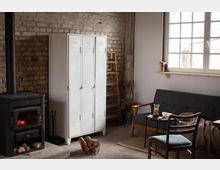 Gemütlicher Wohnraum mit rustikalem Stil: eine Backsteinwand, ein schwarzer Holzofen mit Holzkorb, daneben ein weißer Metallschrank, eine Holzleiter und ein graues Sofa unter einem großen Fenster mit Sprossen. Ein kleiner Tisch sowie Holzstühle ergänzen die Einrichtung.