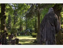 Friedhof mit großen alten Bäumen und Gräbern, umgeben von viel Grün. Im Vordergrund steht die Rückansicht einer Steinstatue, die eine menschliche Figur darstellt.