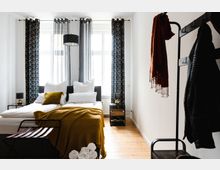 Schlafzimmer mit einem Doppelbett, dekoriert mit weißen Kissen und einer senfgelben Decke, flankiert von zwei Nachttischen mit Lampen. Große, helle Fenster mit gemusterten Vorhängen und ein Kleiderständer mit Jacken und einem Schal an der rechten Seite; Holzboden und minimalistische Einrichtung.